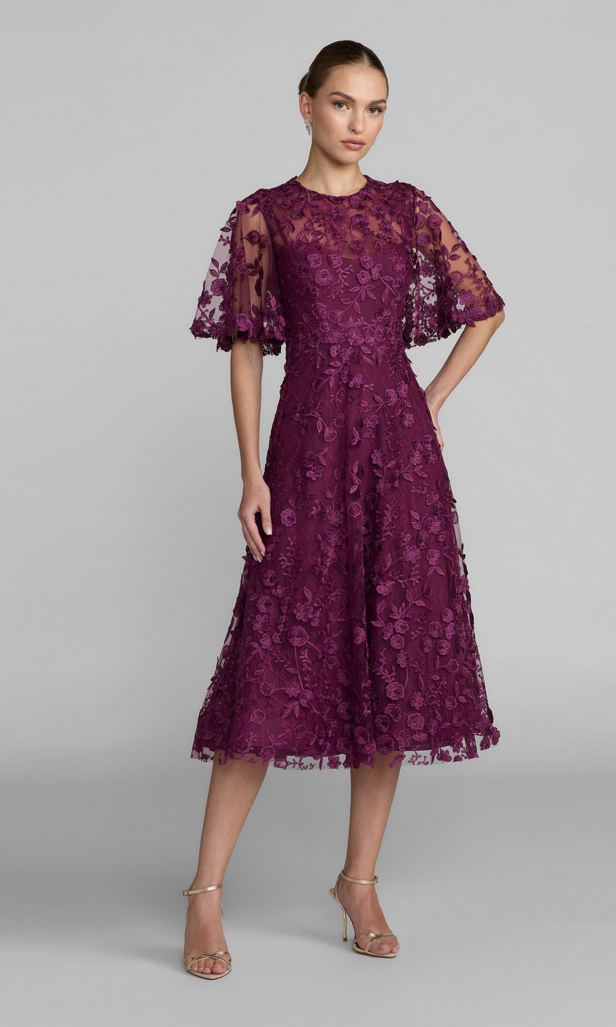 A woman models the Lace Midi Wedding Guest Dress: Mac Duggal 69036, featuring short sleeves, floral lace embroidery, and strappy heels against a plain gray background—a refined pick for wedding guests.